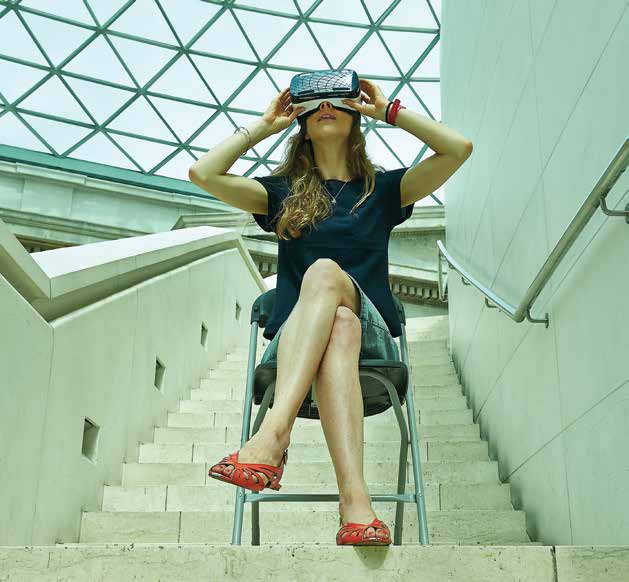 A woman sits on a metal folding chair on a staircase landing with a geometric patterned glass roof above wearing a pair of virtual reality glasses