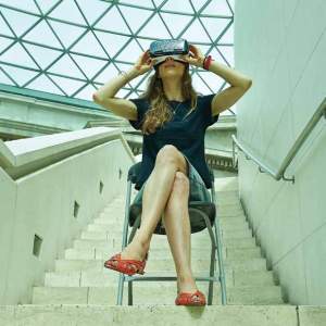 A woman sits on a metal folding chair on a staircase landing with a geometric patterned glass roof above wearing a pair of virtual reality glasses