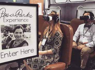 Two visitors sit on bus seats wearing virtual reality glasses with a sign in the foreground saying "Rosa Parks Experience Enter Here"