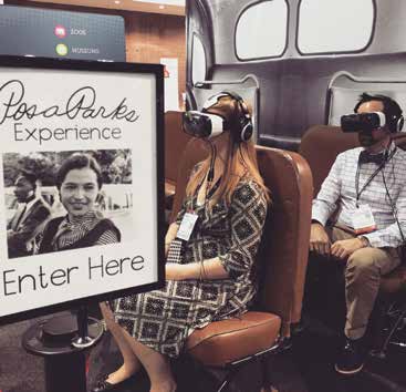 Two visitors sit on bus seats wearing virtual reality glasses with a sign in the foreground saying "Rosa Parks Experience Enter Here"