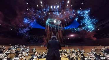 An orchestra plays with the conductor in teh foreground and a light show in the background
