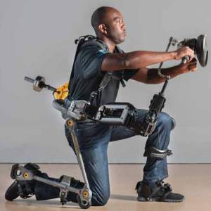 A man kneels on one knee and holds his arms out in front of him wearing an exoskeleton enabling him to do so.