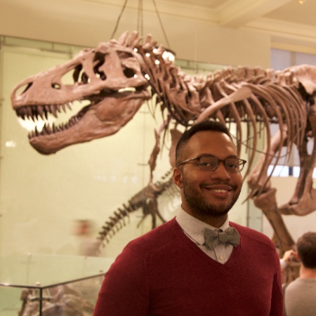 A man stands in front of a T-Rex skeleton wearing a red sweater, white shirt, with a blue or grey bowtie