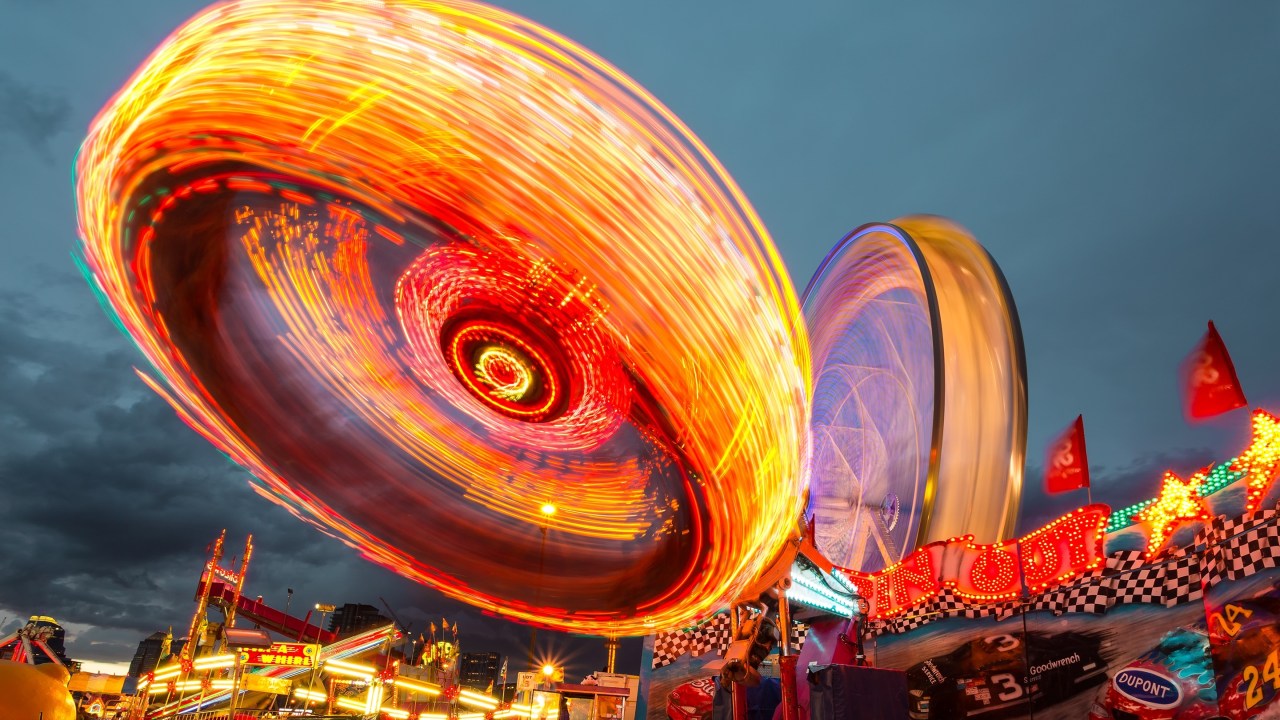 Image of an amusement park ride taken at night. Lights are blurring together to show motion and rides are lit brightly red, orange, and burgundy.