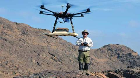 A man stands on a rocky hillside controlling a large drone in the foreground