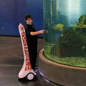 A young man stands next to an aquarium tank with a telepresence robot designed to with giraffe spots