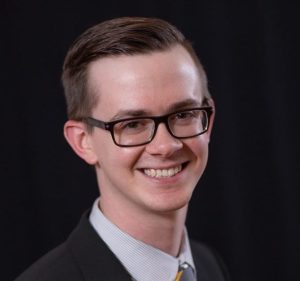 A man stands smiling at the camera wearing a suit and tie and glasses.