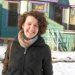 Woman stands in snow wearing heavy winter coat with scarf and short curly brown hair smiling, eyes squinted.