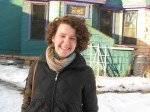 Woman stands in snow wearing heavy winter coat with scarf and short curly brown hair smiling, eyes squinted.