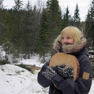 A woman stands in the snow wearing a winter coat and clutching a piece of wood