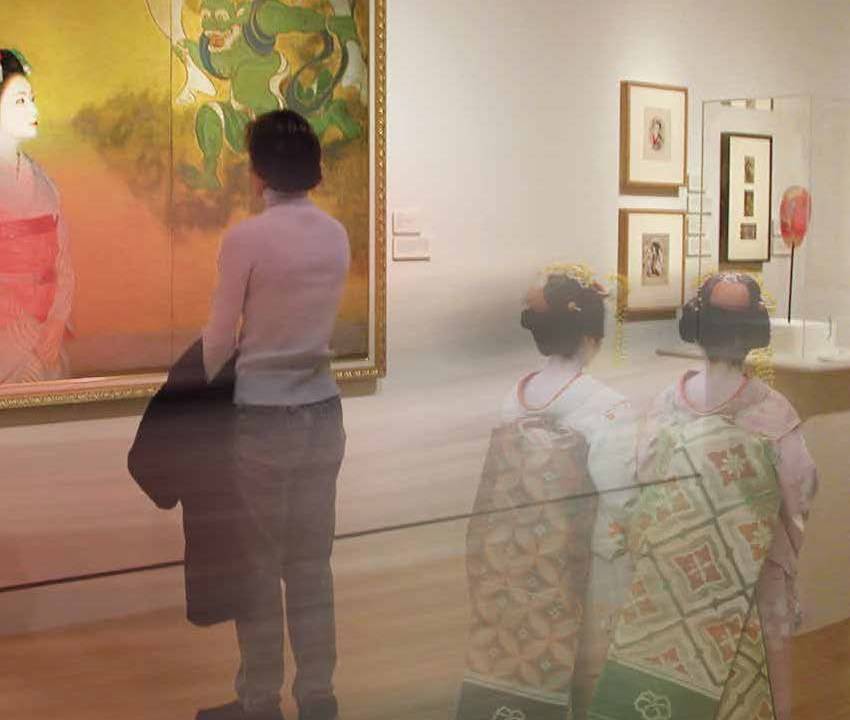 A man stands looking at an Asian artwork while two women dressed in traditional Geisha garb walk behind him