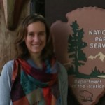 Woman stands in front of a National Park Service sign wearing a multi-colored scarf.