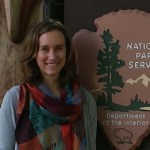 Woman stands in front of a National Park Service sign wearing a multi-colored scarf.