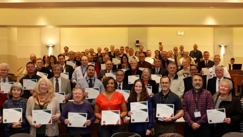 A group of people stand in an auditorium holding up certificates