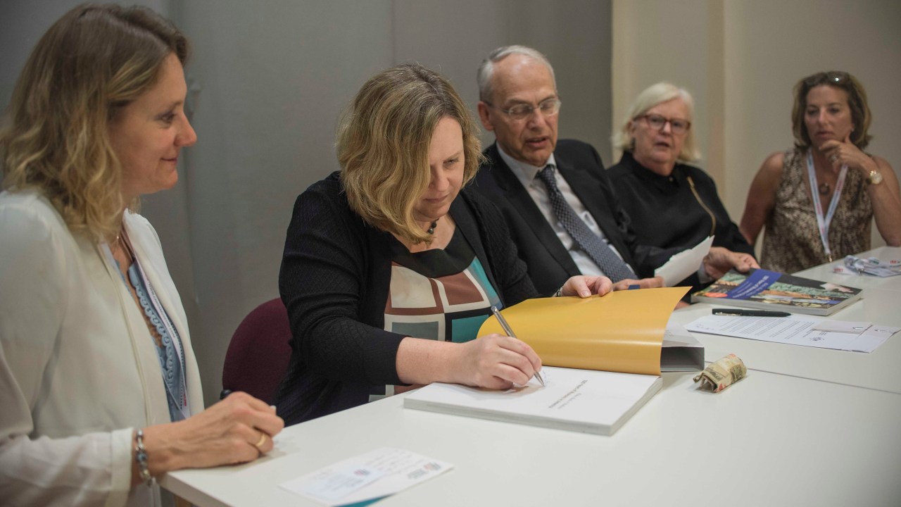 A group of people sit at a long table. Laura Lott is signing the document.