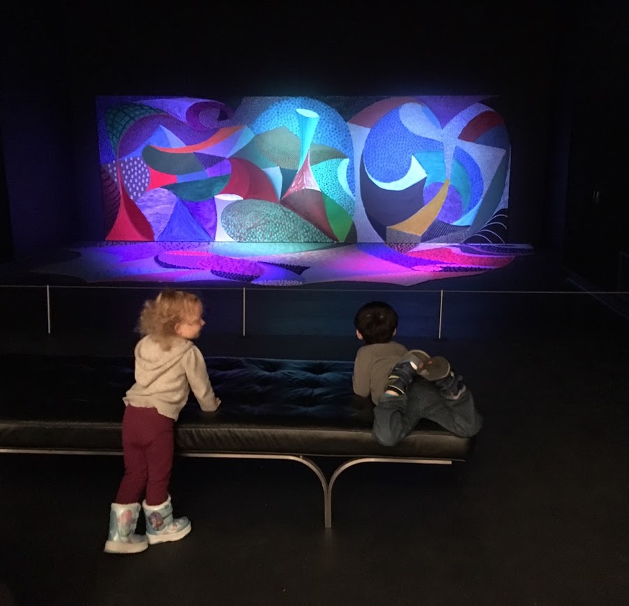 Two children play on a bench in front of a colorful piece of artwork.