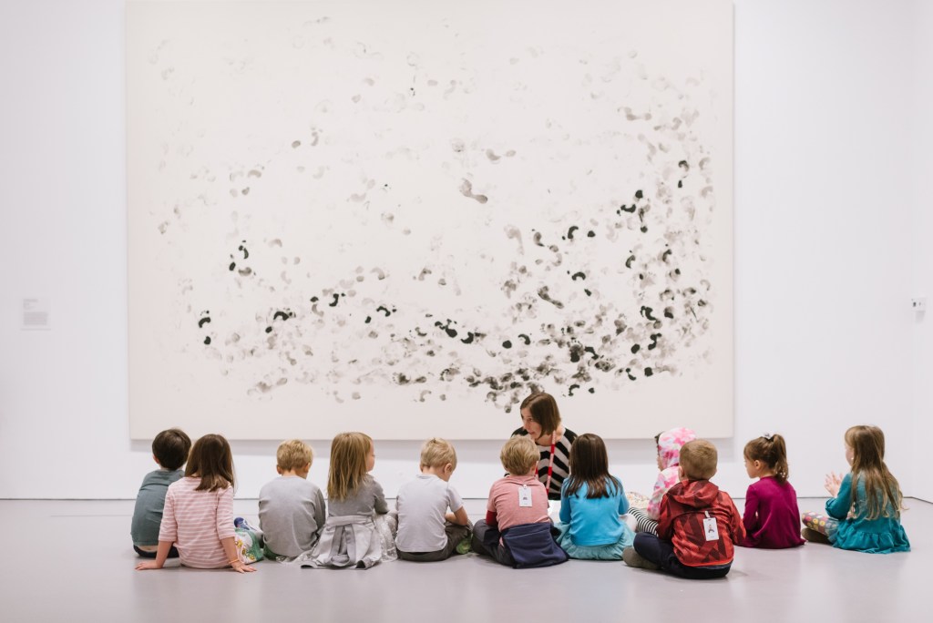 A group of students sit in front of a large artwork listening to a tour guide or teacher.