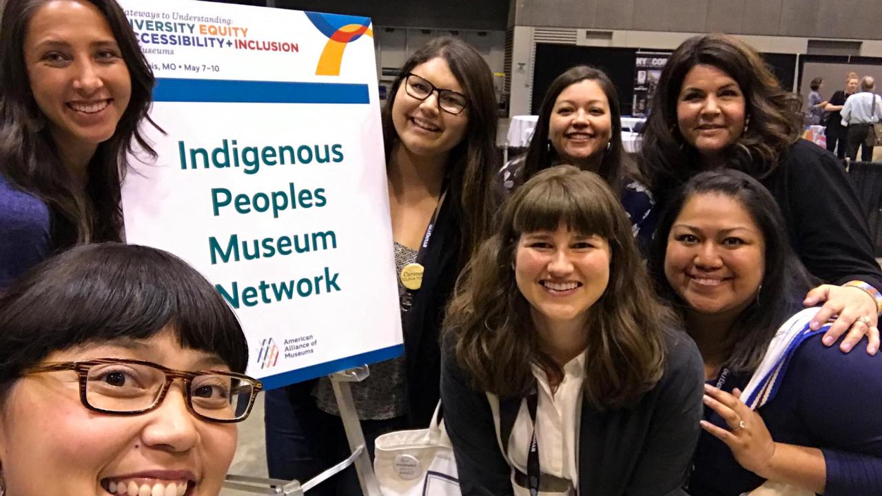 An image of a group of people next to a sign for the Indigenous Peoples Museum Network professional network. They are smiling and standing together.