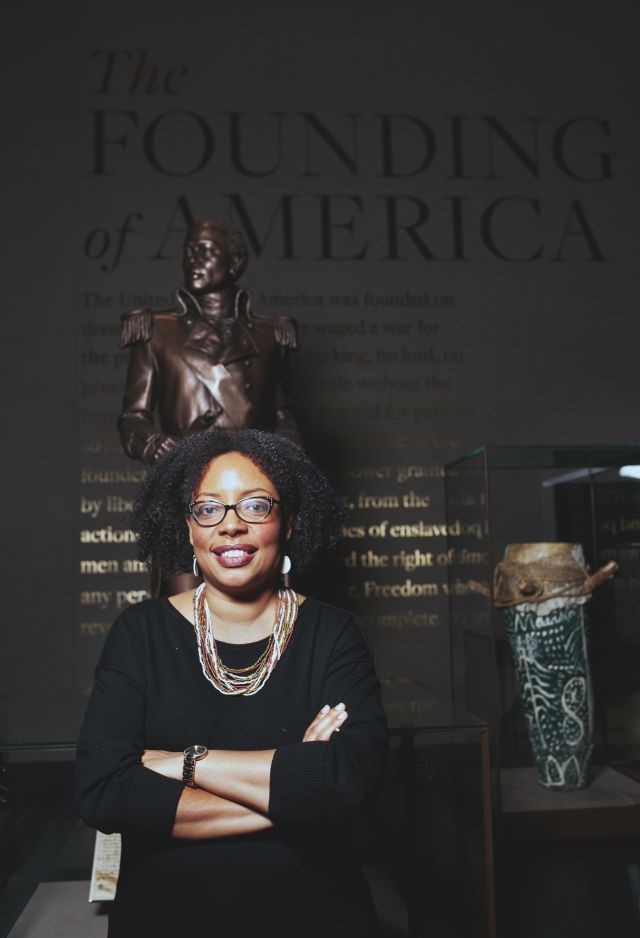 Headshot of Joanne Hyppolite standing in front of a tall sculpture on a dark wall background