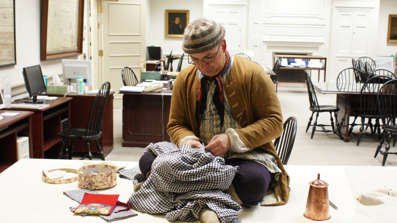 This image features a man seated on a table in a small museum. The man is holding a garment and repairing it by hand. He is wearing a yellow jacket and striped cap.