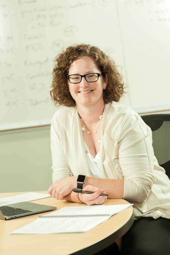 A picture of Sarah Cohn leaning against a desk and smiling at the camera.