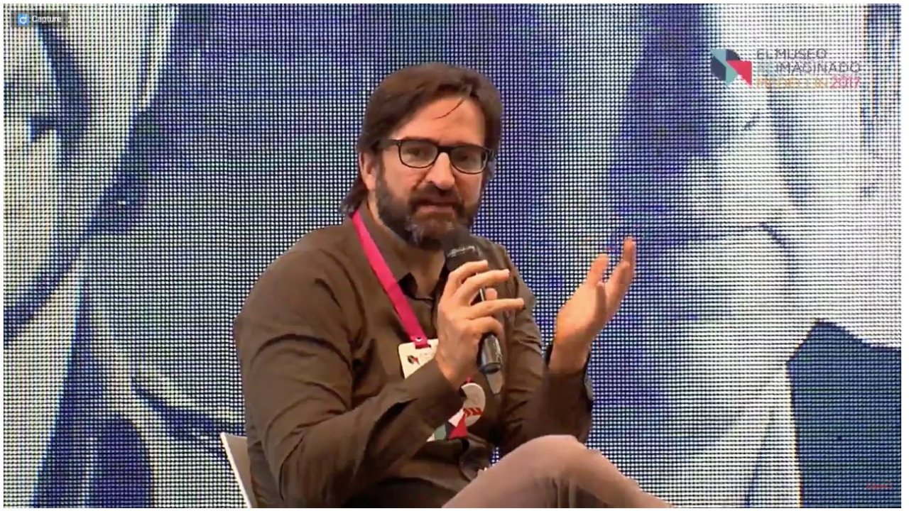 Andres Roldan talking into a microphone and sitting in front of a large video projection