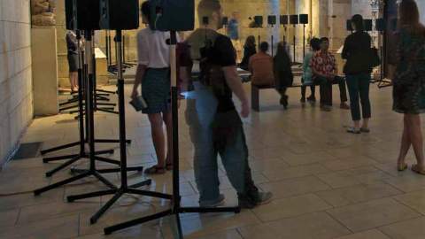 People milling around in a large highly decorative room with speakers at various intervals set up in a circle.