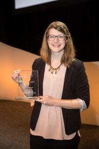 Image of a young woman standing holding a clear glass award while smiling at the camera.