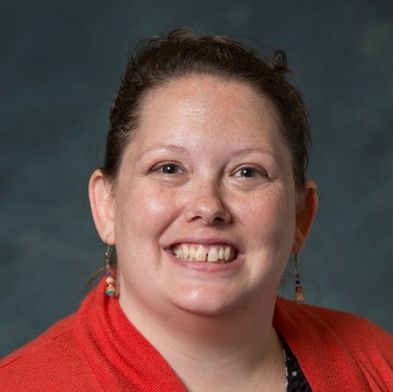 A woman smiles at the camera in front of a grey backdrop wearing her hair up, dangly earrings, a red jacket and black shirt.