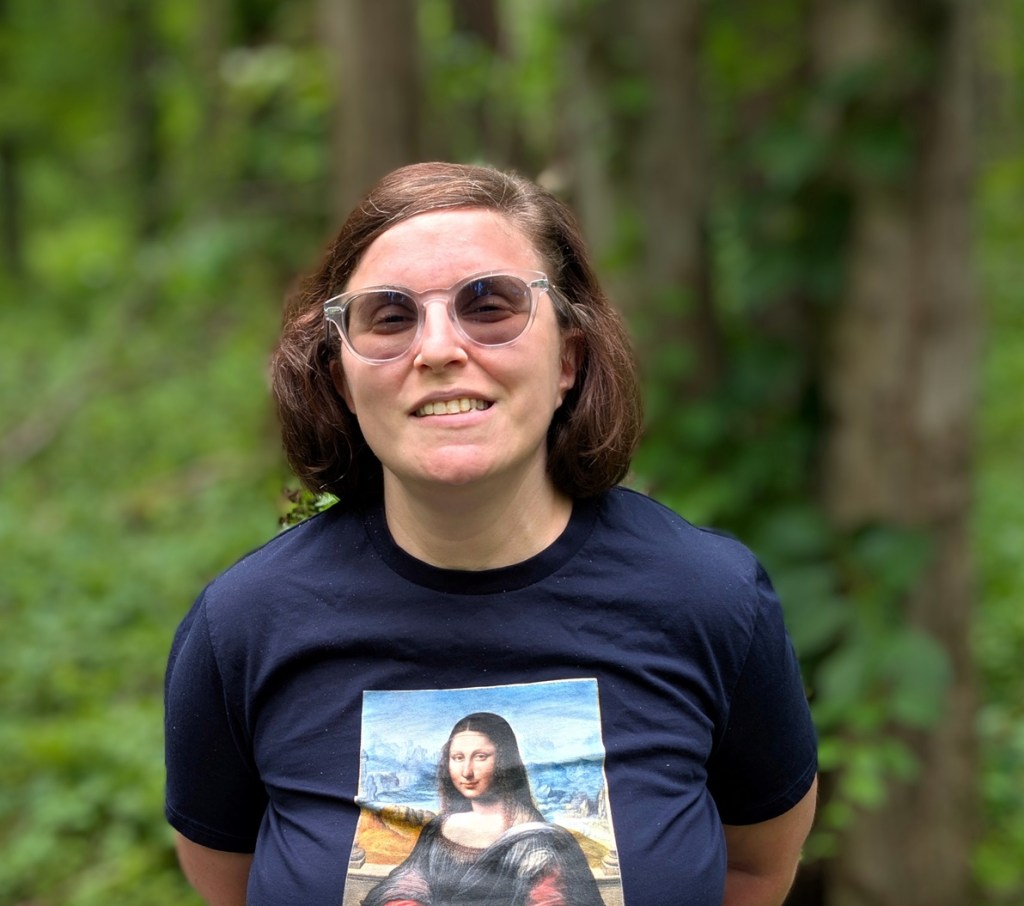Headshot of Aviva Westheim. A person with shoulder length straight brown hair, wearing dark colored glasses and a dark blue t-shirt with a figure on it. 