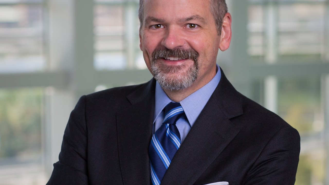 A man stands with his hands folded smiling at the camera wearing a dark suit with a blue shirt and blue striped tie.