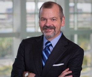 A man stands with his hands folded smiling at the camera wearing a dark suit with a blue shirt and blue striped tie.