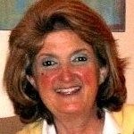 A woman smiles at the camera with shoulder length reddish brown hair wearing a white shirt and yellow sweater.