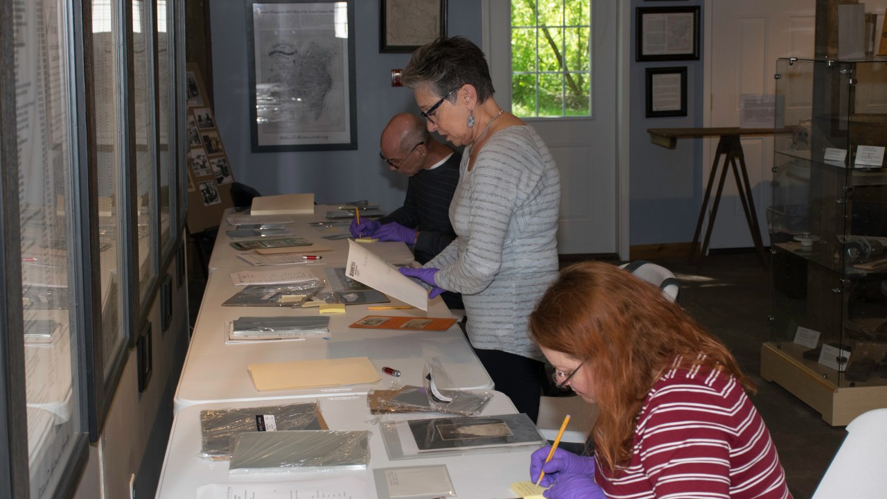 A photo showing three people seated at a table, working with original collections. All have gloves on and are writing notes or looking at the objects.