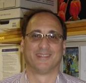 A man stands smiling at the camera wearing glasses and a striped shirt.