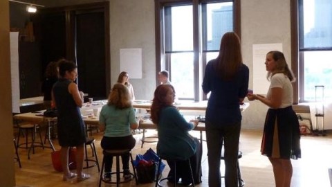 people are seated around a conference table with sunlit windows in the background. Two people stand talking to each other in the foreground