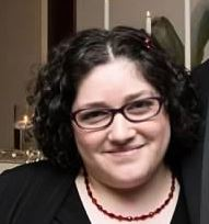 A woman smiles at the camera with short curly dark hair, wearing glasses a dark top and a red necklace.