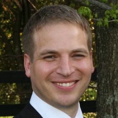 A man smiles at the camera with short blond/brown hair wearing a suit with a white shirt.