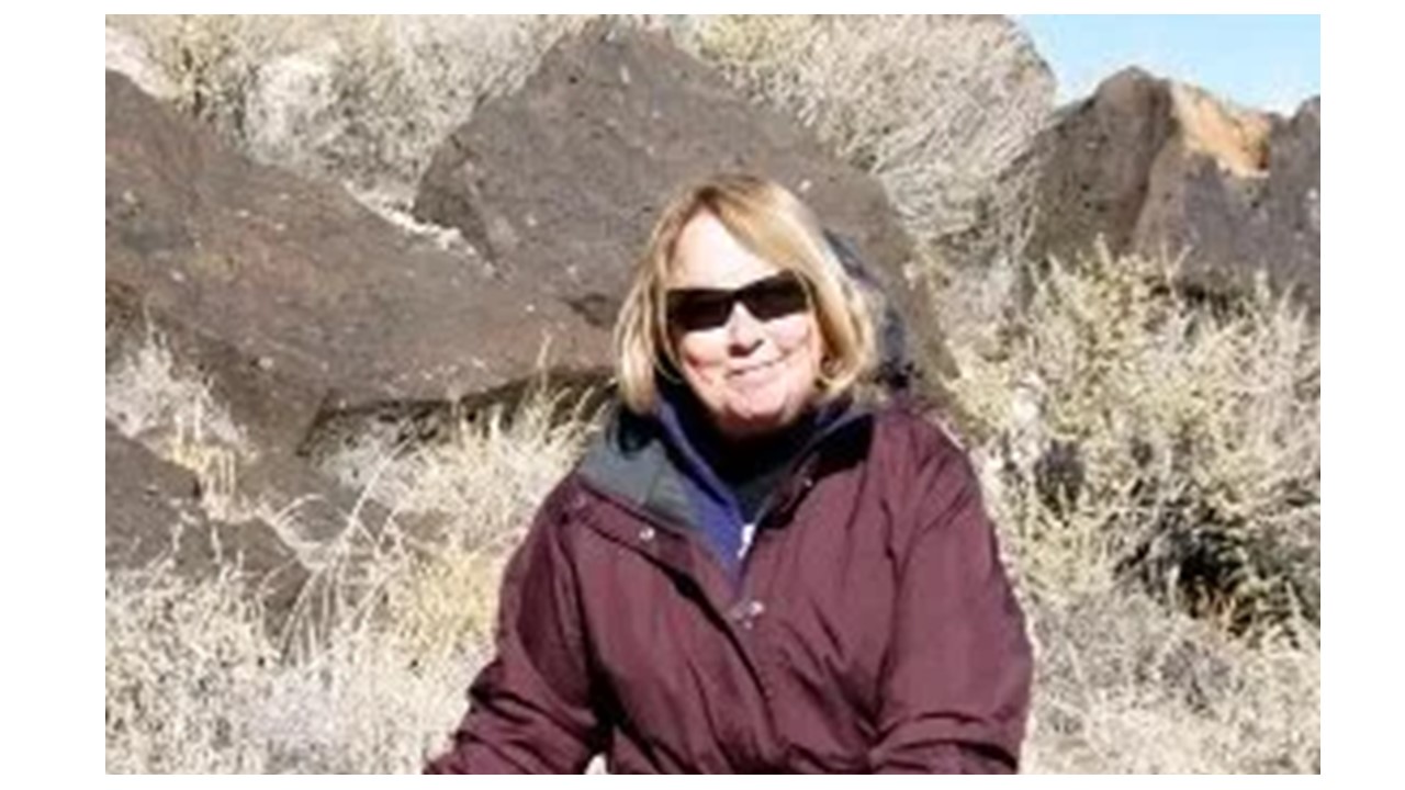 A white woman with blond hair sits leaning against a rock wearing a purple jacket wearing sunglasses.