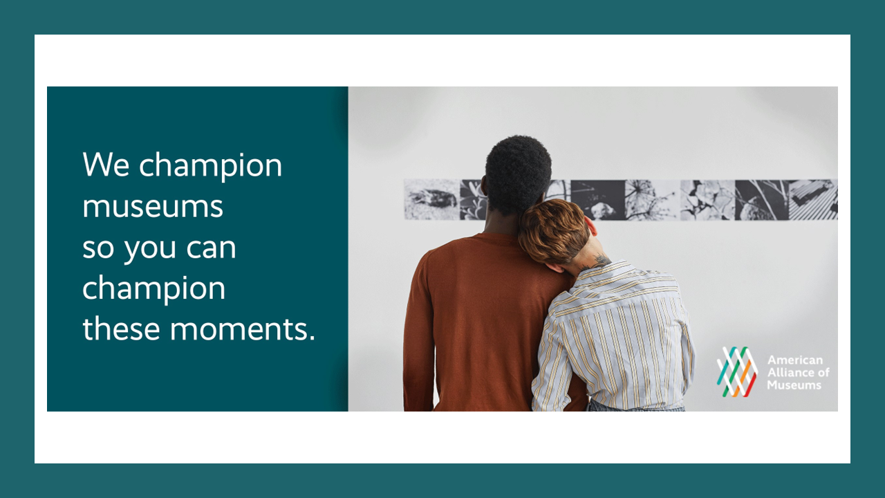 One person leans their head against another taller person's shoulder as they gaze at an exhibition wall with a series of black and white images. Next to them text reads "We champion museums so you can champion these moments."