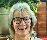 Image of woman smiling at camera in front of a brick background. She has shoulder length hair and is wearing glasses with a gray shirt on. 