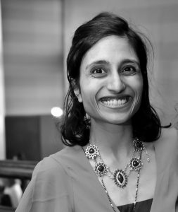 A black and white image of a woman standing slightly off-center smiling with dark hair wearing an elaborate necklace.