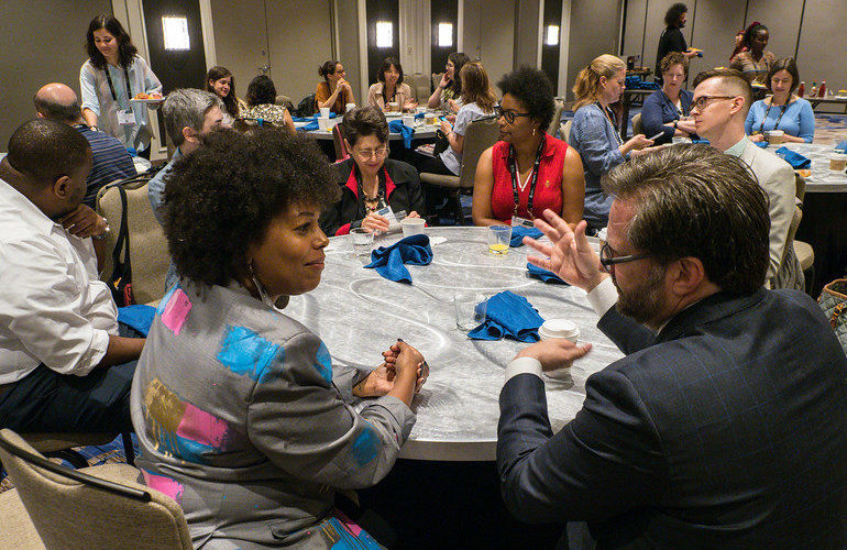 Several people sit around a round table discussing topics on blue cards in front of them.