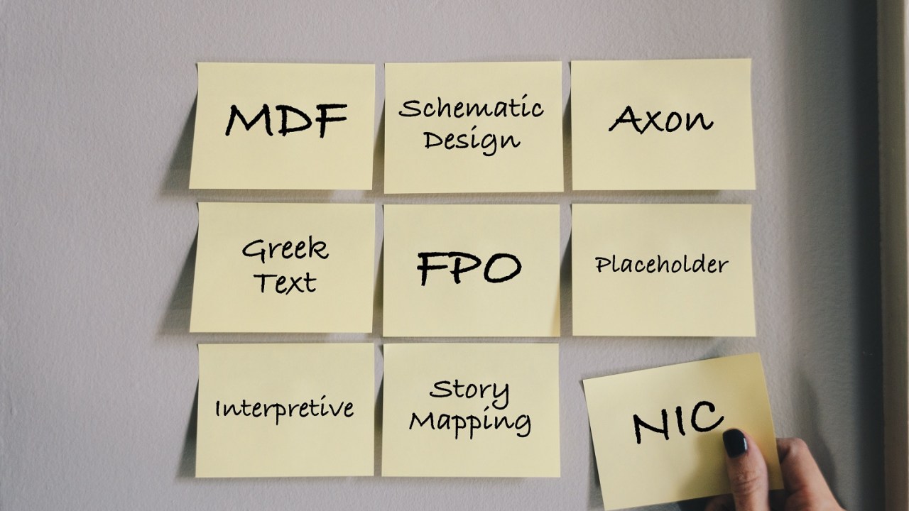 Image of 12 Post-It notes on a wall with various acronyms or design words. Top row: MDF, Schematic Design, Axon. Second row: Greek Text, FPO, Placeholder. Third row: Interpretive, Story Mapping, NIC. The final note is being held by a woman's hand wearing dark nail polish.