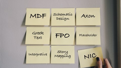 Image of 12 Post-It notes on a wall with various acronyms or design words. Top row: MDF, Schematic Design, Axon. Second row: Greek Text, FPO, Placeholder. Third row: Interpretive, Story Mapping, NIC. The final note is being held by a woman's hand wearing dark nail polish.