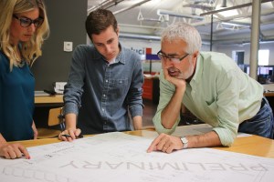 Three people leaning over a desk reviewing a large piece of pager during a design review session.