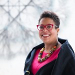 A woman smiles at the camera with short cut dark curly hair with red highlights wearing red rimmed glasses, a bright pink top, with a dark shrug jacket, and a brightly colored chunky necklace.