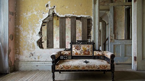 Image of a wall with the plaster partially removed showing the lathe and wooden studs beneath with a new couch sitting just into a doorway into another room.