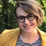 A woman smiles at the camera wearing glasses with short blond hair swept to the left of her face. She is wearing a black and white striped top with a yellow jacket.