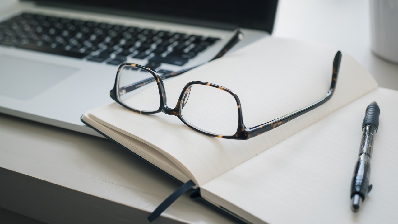 Image of a laptop blurred in the background with an open book and a pair of horn rimmed glasses sitting on the book.